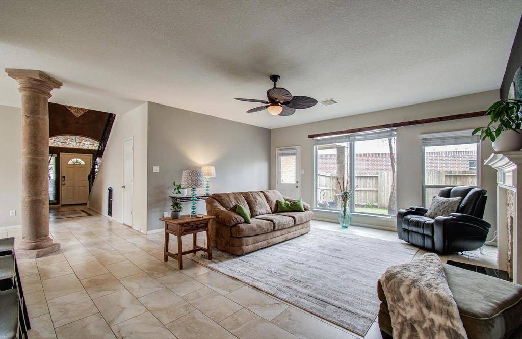 Spacious living room with large windows, offering plenty of natural light. Features include tiled floors, a ceiling fan, and a cozy seating area. The room is connected to the entrance hallway, accented with a decorative column and views of the fenced backyard.