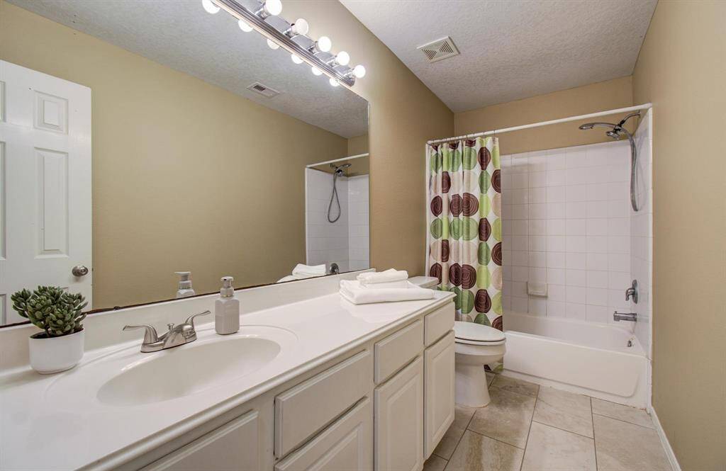 This bathroom features a spacious vanity with dual sinks, modern fixtures, and ample storage. It includes a shower-tub combo with a colorful curtain, and bright lighting enhances the clean, neutral decor.