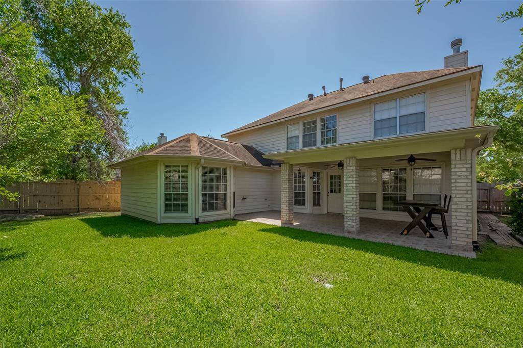 Well maintained backyard, with a spacious covered patio, featuring double  ceiling fans and a dining area. The yard is grassy and fenced, surrounded by mature trees, offering privacy and a serene outdoor space.