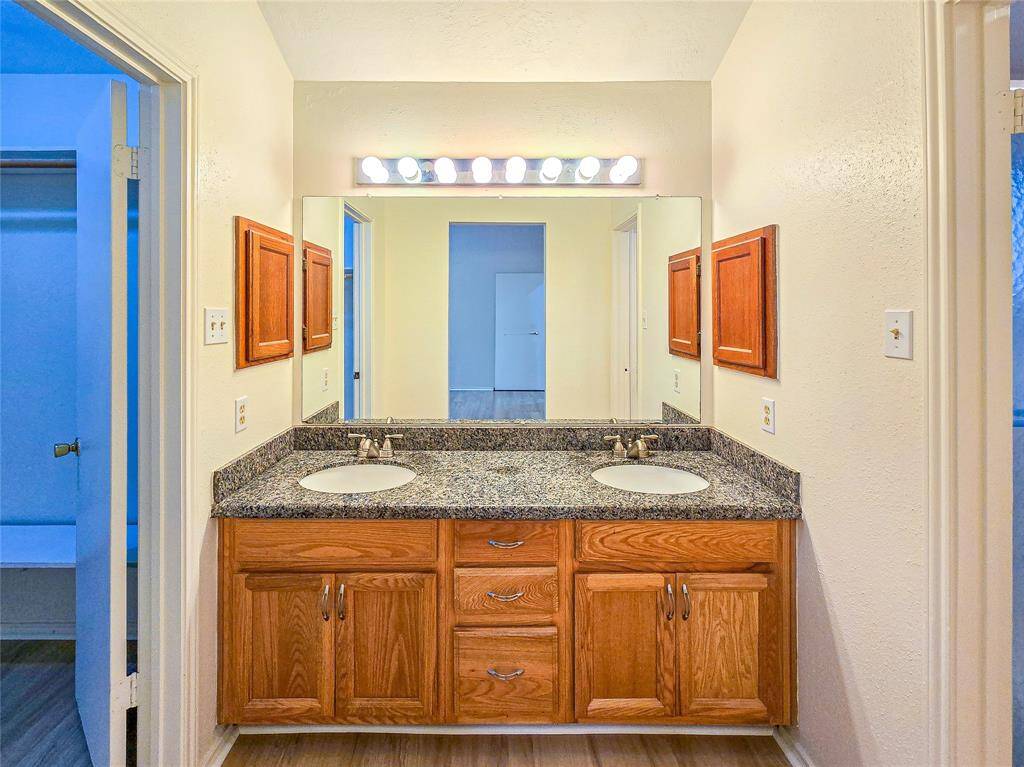 Bathroom features a dual-sink vanity with granite countertops and ample storage. It includes classic wooden cabinets and a large mirror under bright lighting, offering a functional and stylish space.