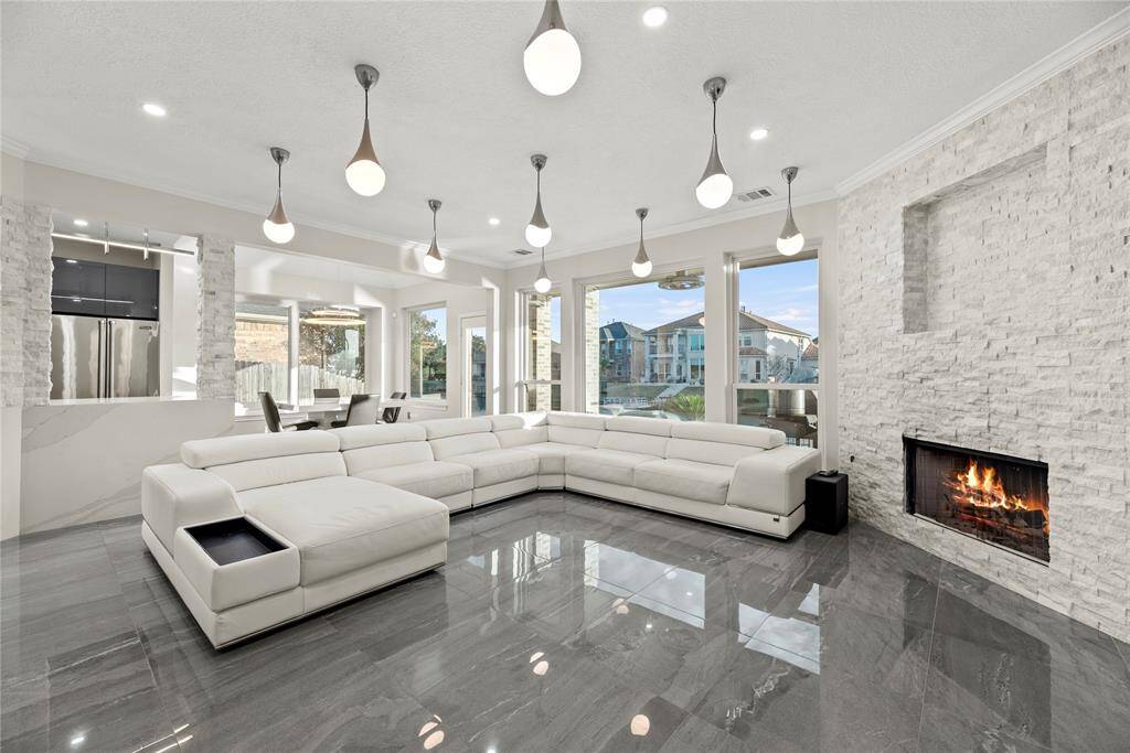 Luxurious open-concept family room featuring a modern fireplace, elegant lighting, expansive windows with serene water views, and sleek tile floors, perfect for both relaxation and entertaining.