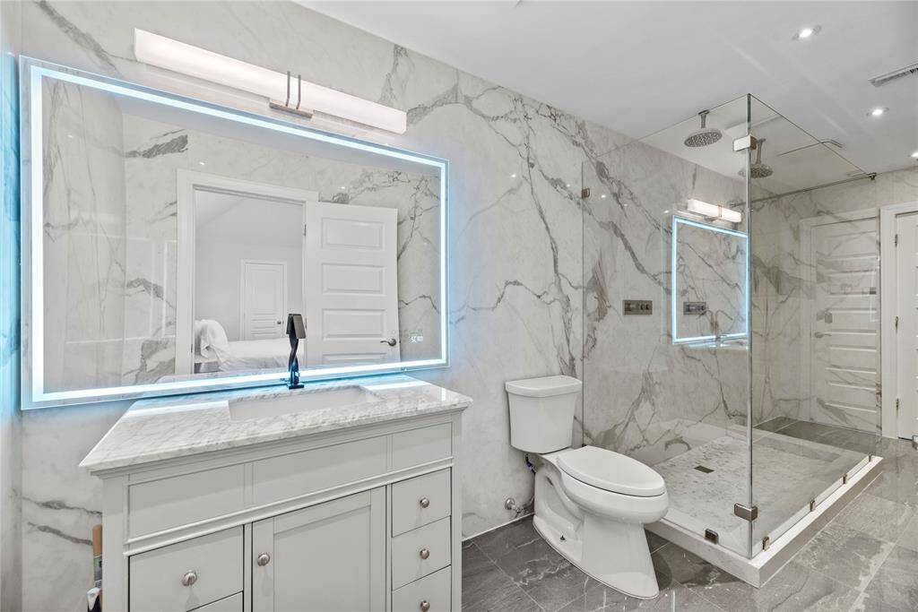 Luxurious upstairs bathroom featuring sleek marble finishes, an illuminated vanity mirror, and a spacious glass-enclosed rainfall shower.