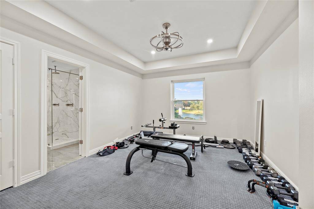 Upstairs bedroom currently set up as a home gym, featuring plush carpeting, tray ceiling with modern lighting, and a window offering serene water views, adjacent to a luxurious ensuite bathroom.