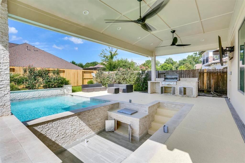 Beautiful outdoor space featuring a pool and a covered patio. The pool has a modern design with clean lines and a light-colored stone surround. The patio area is spacious, equipped with a built-in grill and countertop for outdoor cooking. There are ceiling fans installed for comfort, and a mounted TV for entertainment.