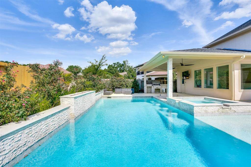 Comfortable outdoor seating is arranged near the pool, and the area is well-landscaped with bushes and plants along the fence line, providing privacy and a pleasant atmosphere. The overall setting is perfect for relaxation and outdoor gatherings.