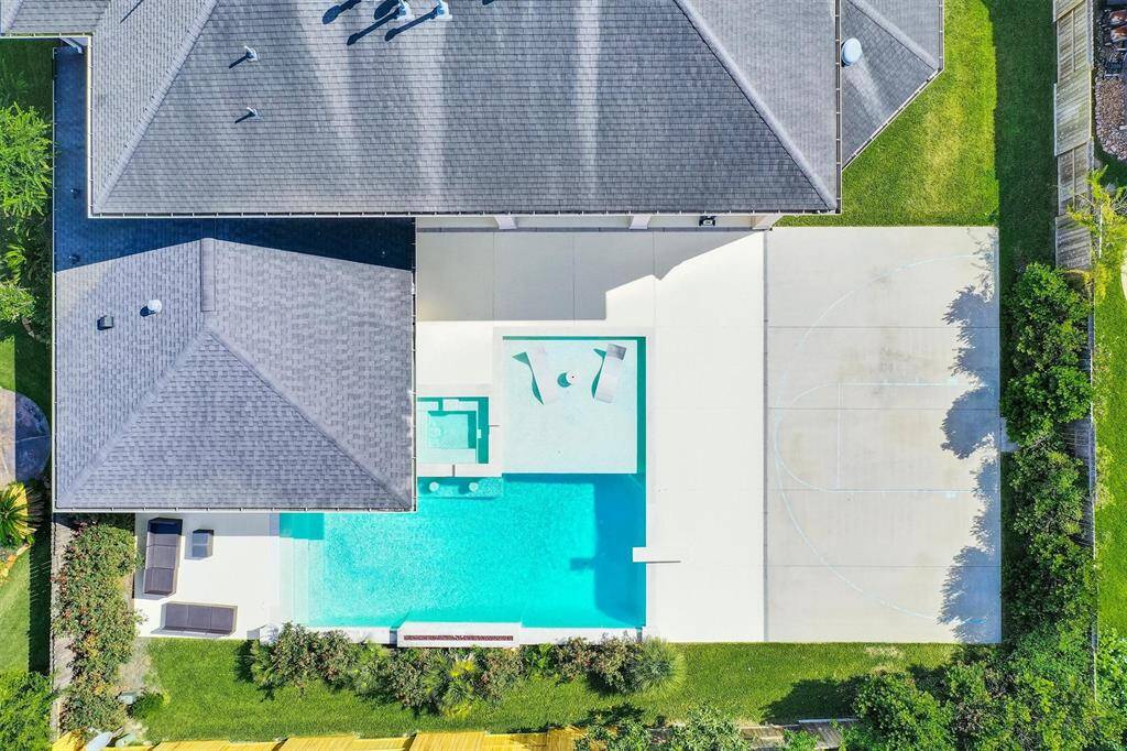 An aerial view of this beautiful outdoor space featuring a pool and a covered patio. The pool has a modern design with clean lines and a light-colored stone surround. The patio area is spacious, equipped with a built-in grill and countertop for outdoor cooking. There are ceiling fans installed for comfort, and a mounted TV for entertainment.