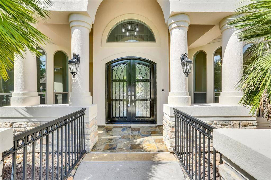 A close up shot of the entrance of the entry way showcasing beautiful railings, custom tile flooring, gorgeous french doors, arched walkways, beautiful landscaping and covered front porch.