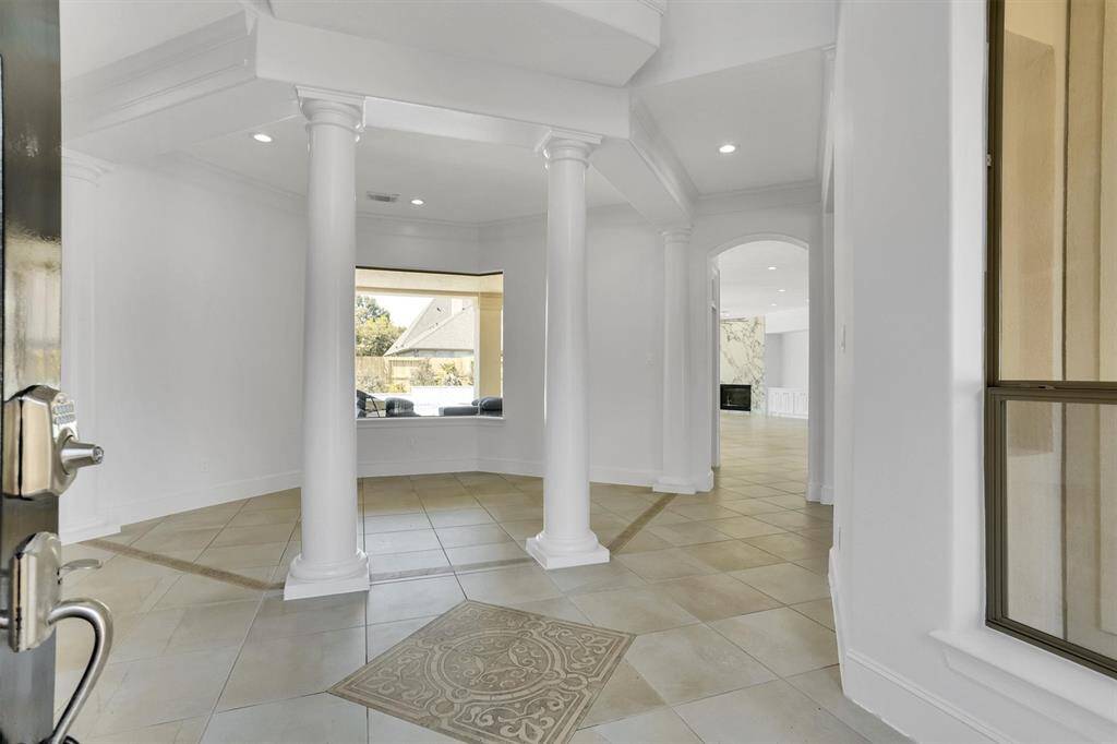 As you step inside, you are greeted by a spacious foyer with a high ceiling, highlighted by two prominent white columns. The flooring consists of large, light-colored tiles with a decorative tile inset near the entrance. The walls are painted white, contributing to the bright and open feel of the space.
