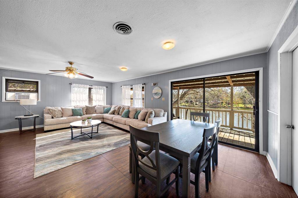 Spacious living area with a comfortable sectional sofa, dining table, and sliding glass doors leading to a deck with a scenic water view. The room features wood flooring, light blue walls, and abundant natural light.