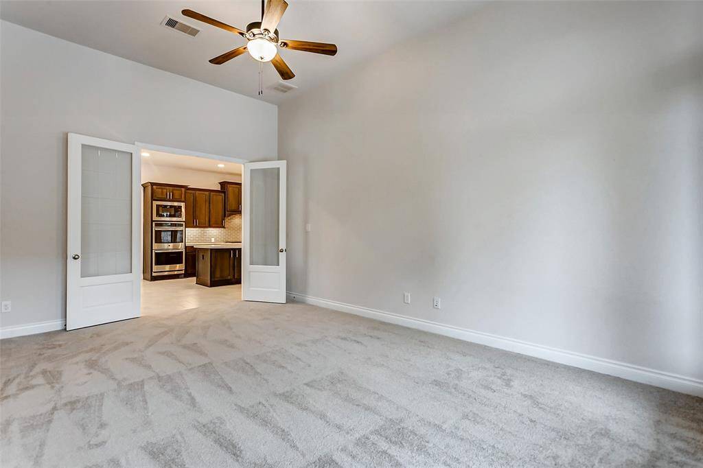 The elegant design of the French doors introduces a touch of sophistication while allowing natural light to flood the room, creating a bright and airy atmosphere. A soaring ceiling with a ceiling fan, coupled with plush carpeting, adds to the comfort and warmth of the space. Large windows along the back wall further enhance the feeling of openness.