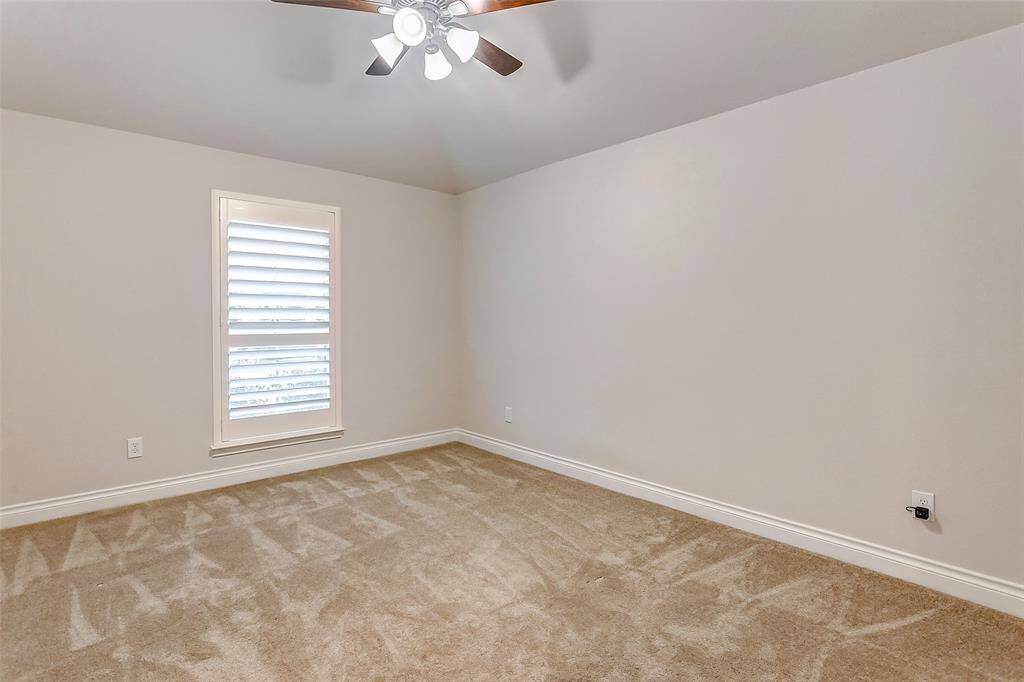 This secondary bedroom features a high ceiling with a ceiling fan, adding to the open, airy feel of the space. Plush carpeting provides warmth and comfort, while a large window, equipped with 2-inch blinds, allows abundant natural light to brighten the room, creating a welcoming and relaxing environment.