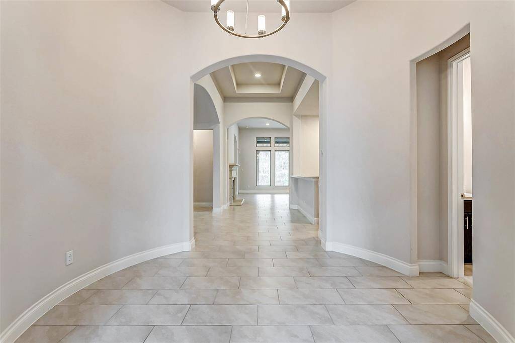 The foyer gracefully leads into a rotunda-like space, where an elegant chandelier hangs overhead, casting a soft, inviting glow. The elongated tile floors flow seamlessly throughout the main living areas, adding a touch of sophistication and continuity to the home’s design.