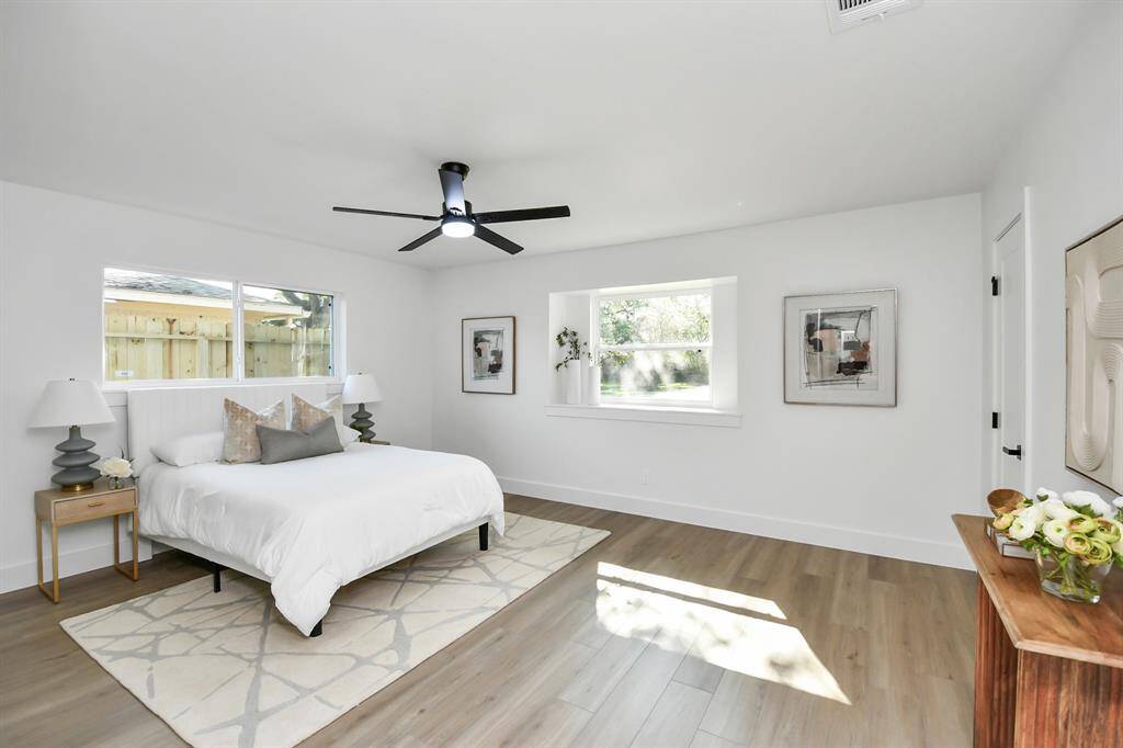 This is a bright, modern bedroom featuring waterproof rigid core luxury vinyl flooring, a ceiling fan, and ample natural light from two large windows. The room is staged with a bed, side tables, lamps, and a few decorative items, showcasing a clean, minimalist aesthetic.