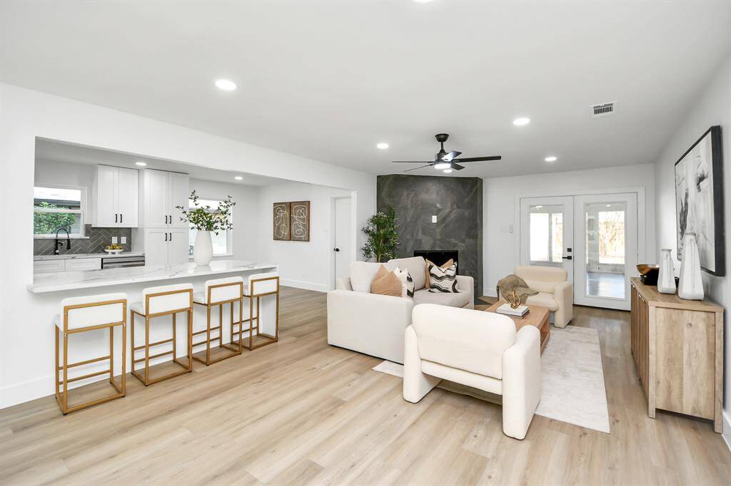 This is a bright and modern open-plan living space featuring a cozy seating area with a fireplace, a dining area visible in the background, and a sleek kitchen with white cabinetry. The room is well-lit with recessed lighting and natural light from large windows, providing a welcoming atmosphere for entertaining and relaxation.