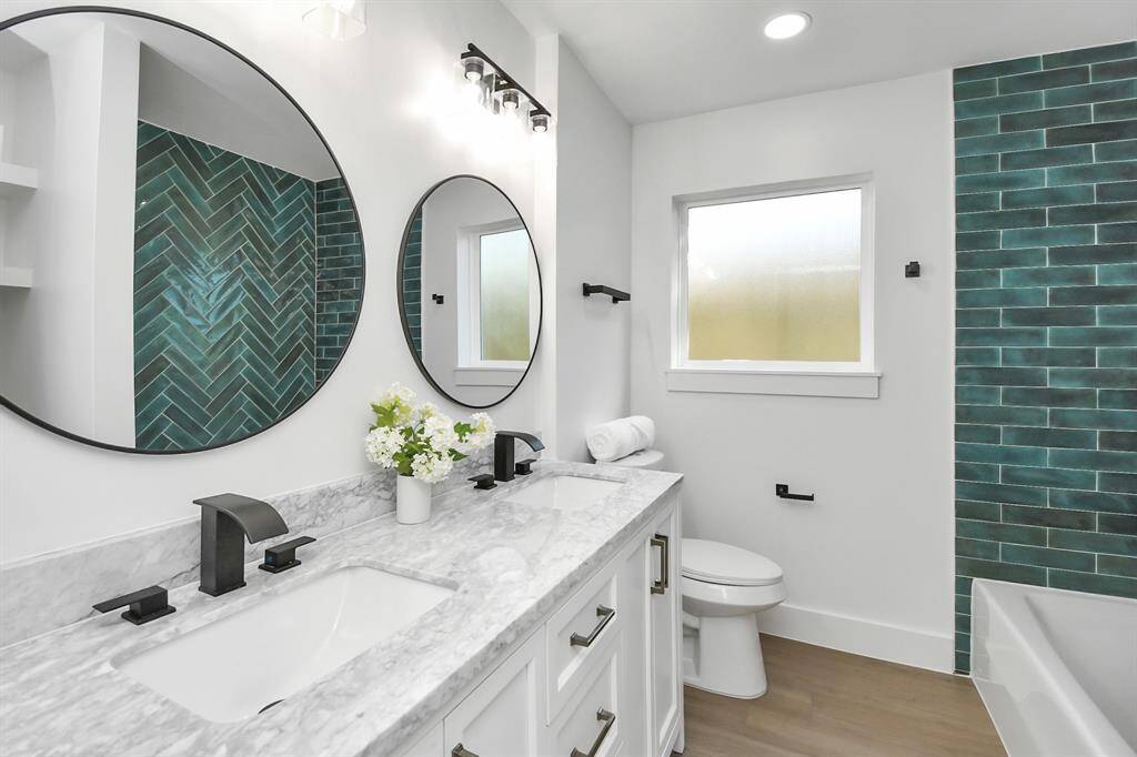 This modern bathroom features a double vanity and sleek black fixtures, a large circular mirror, herringbone-patterned teal tiles in the shower area, and a frosted window for privacy. The design is clean and contemporary with white walls and a wood-look floor.