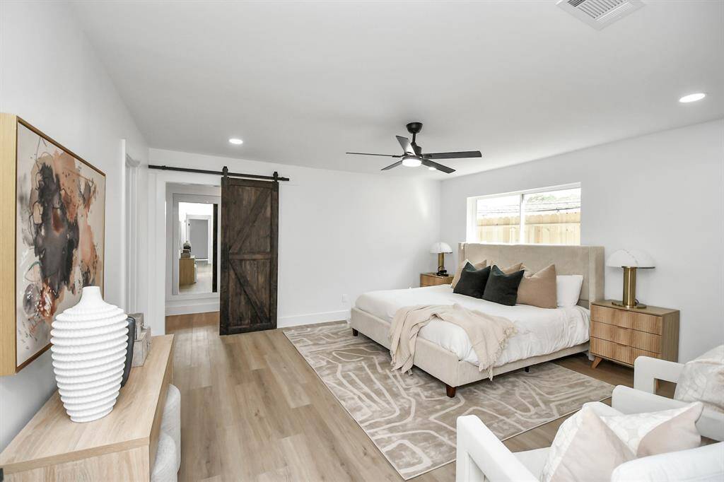 This spacious and well-lit primary bedroom features modern finishes, a ceiling fan, and a sliding barn door leading to a custom walk-in closet. The light waterproof rigid core luxury vinyl flooring and neutral color palette enhance the room’s open and airy feel.
