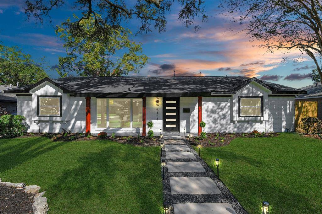 This is a charming, single-story home featuring a white exterior with contrasting dark trim and red accents. It has a neat, landscaped front yard with a pathway leading to the front door and large windows that allow for plenty of natural light. The property is set against a backdrop of a beautiful sunset sky.