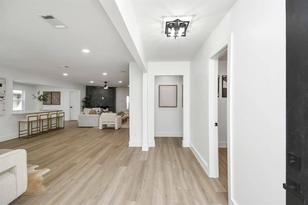 The photo showcases a modern, open-plan interior with a spacious layout, light luxury vinyl flooring, and recessed lighting that creates a bright and welcoming atmosphere. The view leads into a cozy living area, and there's an elegant light fixture as an accent in the corridor.