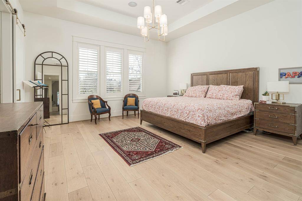Spacious primary bedroom features hardwood floors, modern lighting. Natural light streams through three windows with shutters.