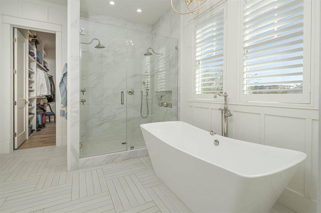 This elegant bathroom features a spacious glass-enclosed shower, a freestanding bathtub, and large windows with plantation shutters for ample natural light.