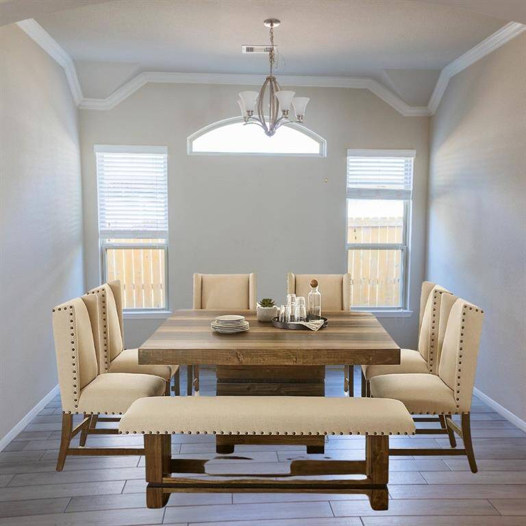 This dining room features a stylish wooden table with upholstered chairs and a bench. Natural light fills the space through two windows, and a modern chandelier adds an elegant touch. The room has a neutral color palette and tiled flooring.
