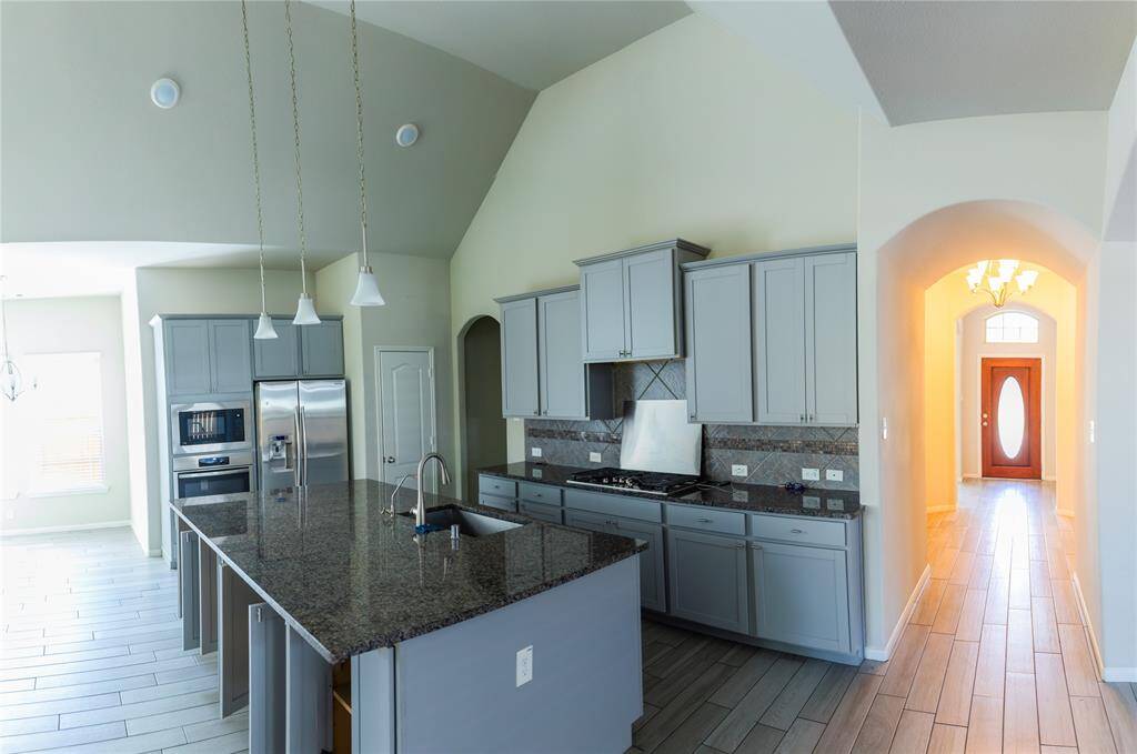 This modern kitchen features high ceilings, granite countertops, stainless steel appliances, and ample cabinetry. The open layout connects to a bright hallway leading to the entrance, creating a welcoming and spacious atmosphere.