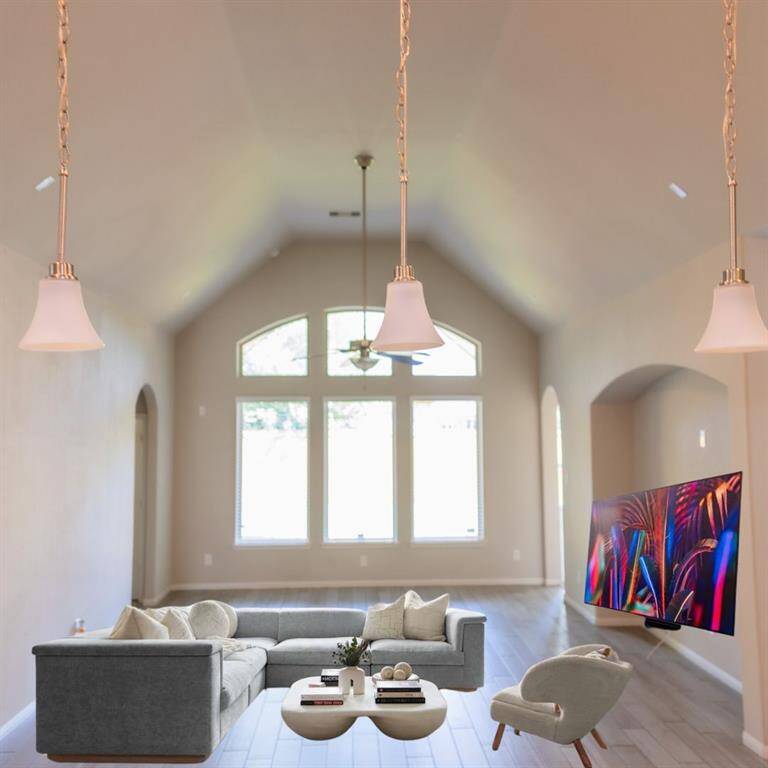 This spacious living room features high ceilings, ample natural light from large windows, modern pendant lighting, and wood flooring. It includes a comfortable gray sectional, stylish armchair, and a wall-mounted TV, creating an inviting space for relaxation and entertainment.