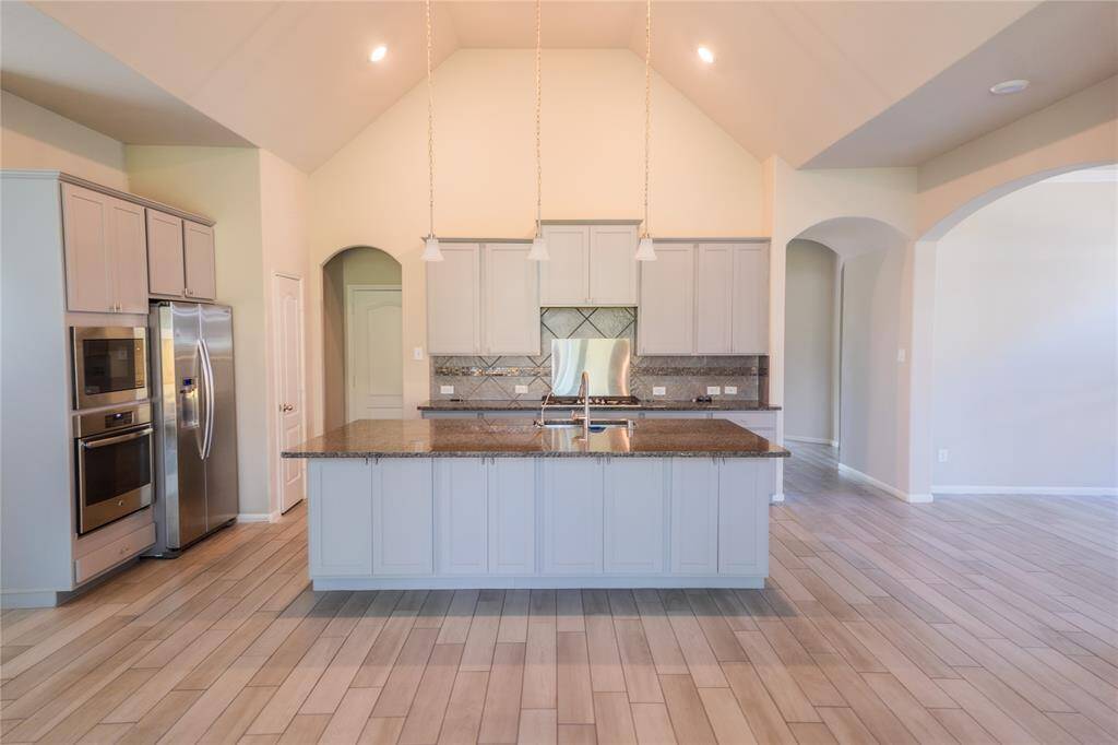 This modern kitchen features high ceilings, a central island with granite countertops, stainless steel appliances, and ample cabinetry. The open layout and light wood flooring create a spacious and inviting atmosphere.
