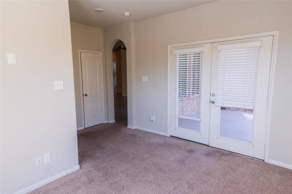 This room features a cozy carpeted floor, neutral walls, and double glass doors with blinds leading to a patio. An arched doorway and a standard door provide access to adjacent spaces, creating a bright and open atmosphere.