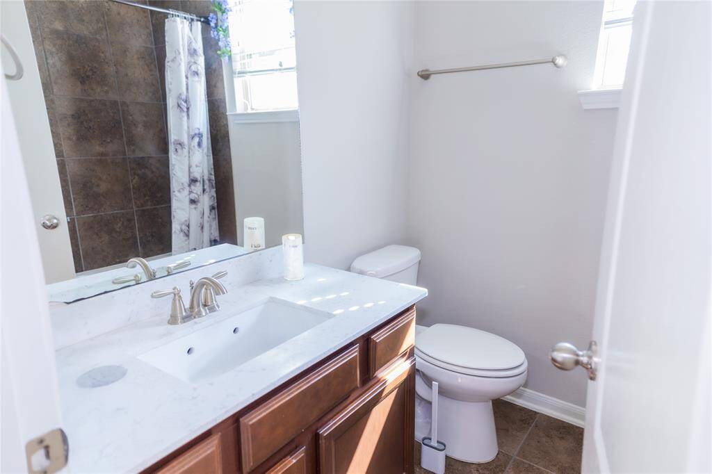 This bathroom features a modern design with a marble countertop, under-mounted sink, and wood cabinetry. It includes a toilet and a shower with tiled walls and a floral curtain, all illuminated by natural light from a small window.