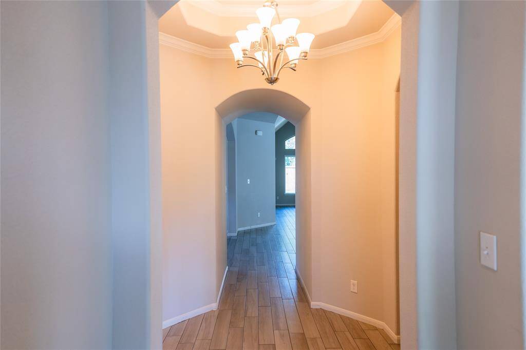 This photo showcases a welcoming hallway with elegant lighting, crown molding, and wood-like tile flooring, leading to a bright room with large windows.