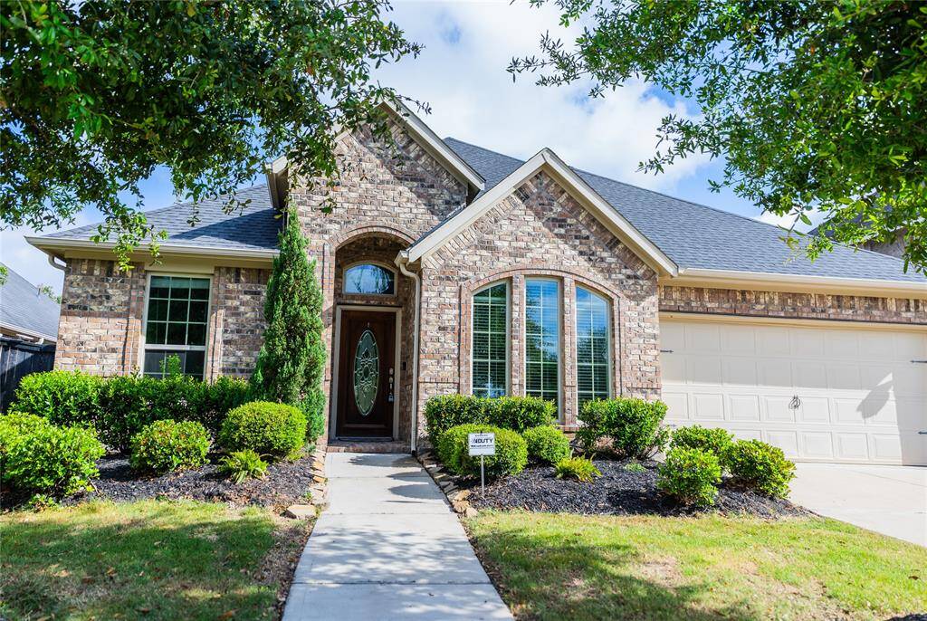 Charming single-story brick home with a gabled roof, featuring a welcoming arched entryway and large front windows. The tidy landscaping includes neatly trimmed bushes and a curved walkway leading to the entrance. The attached 3-car garage offers convenience and ample space.