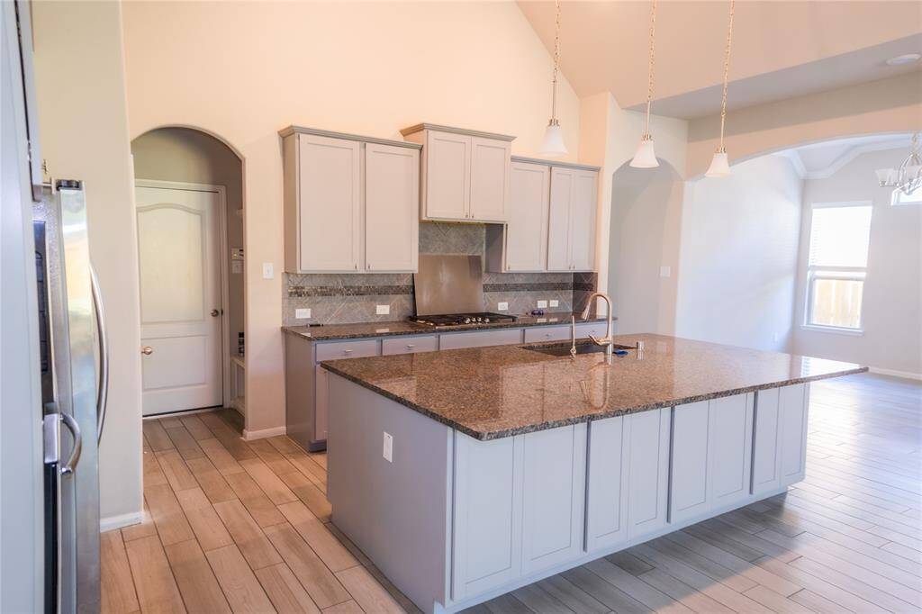 A spacious, modern kitchen with light cabinetry and a large granite island. It features stainless steel appliances, pendant lighting, and wood-look tile flooring, offering a bright and inviting atmosphere.
