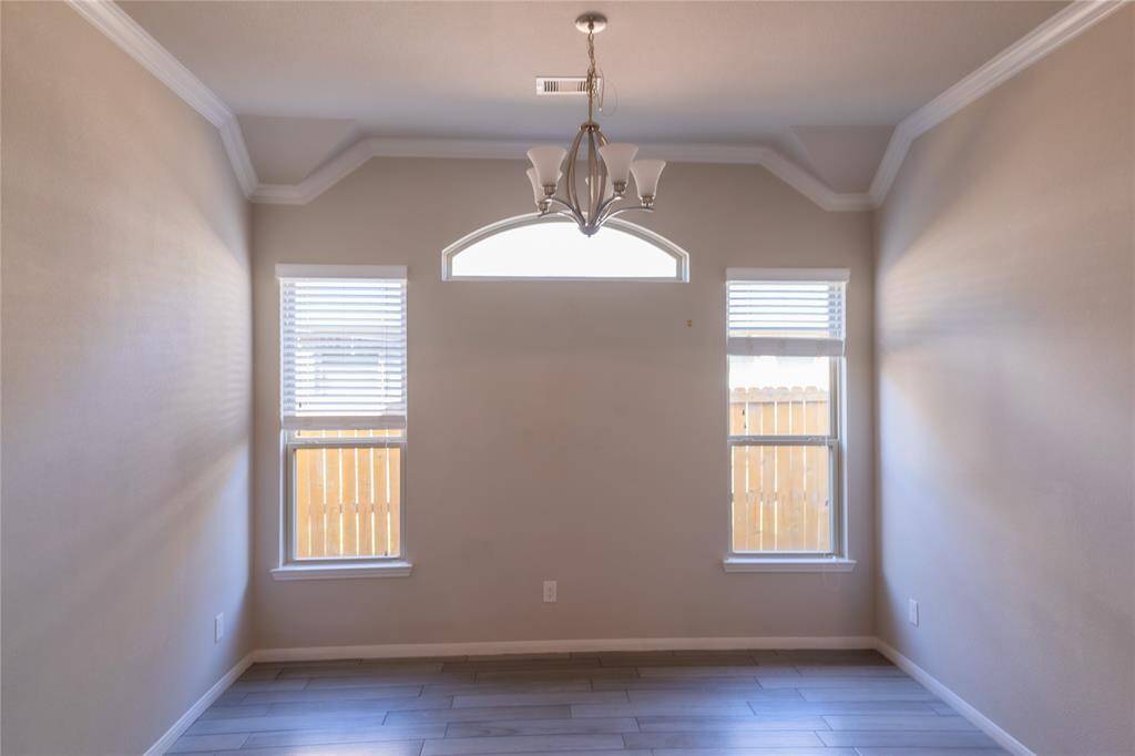 This room features a high ceiling with crown molding, a modern chandelier, and ample natural light from two windows. The neutral walls and wood-look tile flooring create a versatile space, perfect for dining or a home office.