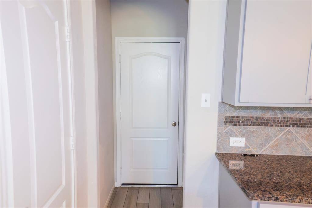 This photo shows a section of a kitchen with granite countertops and tiled backsplash. There’s a white door leading to another room, and the flooring is a warm-toned wood or laminate.