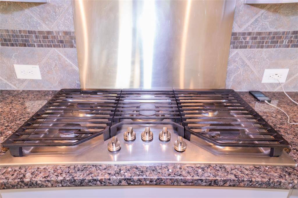 This kitchen features a sleek gas cooktop with five burners set in a granite countertop. The backsplash is a mix of stone and tile, adding a modern touch.