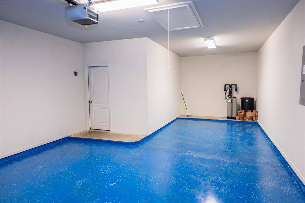 This photo shows a clean, single-car garage with a bright blue epoxy floor, white walls, and ceiling lighting. It includes a closed door, a water treatment system, and some storage space, making it functional and well-maintained.