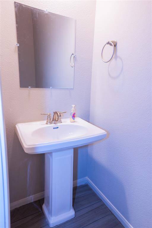 This photo shows a small, well-lit bathroom with a pedestal sink, a wall-mounted mirror, and a towel ring. The space features light-colored walls and wood-style flooring, creating a clean and modern look.