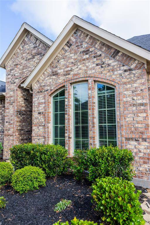 This photo shows a portion of a home exterior featuring a brick facade and a large, arched window with shutters. The surrounding landscaping includes neatly trimmed shrubs and a dark mulch bed, providing an attractive, well-maintained look.