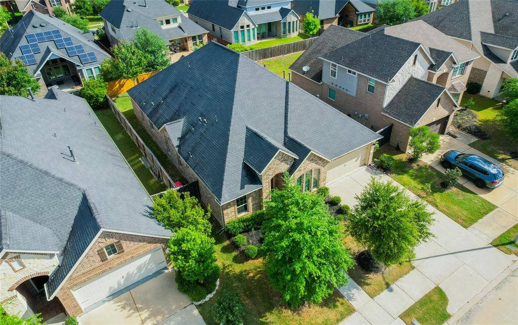 This aerial view showcases a modern, single-family home in a suburban neighborhood. It features a dark roof, brick facade, and a well-maintained front yard with mature trees. The driveway leads to a two-car garage, and the house is surrounded by similar homes, offering a cozy community feel.