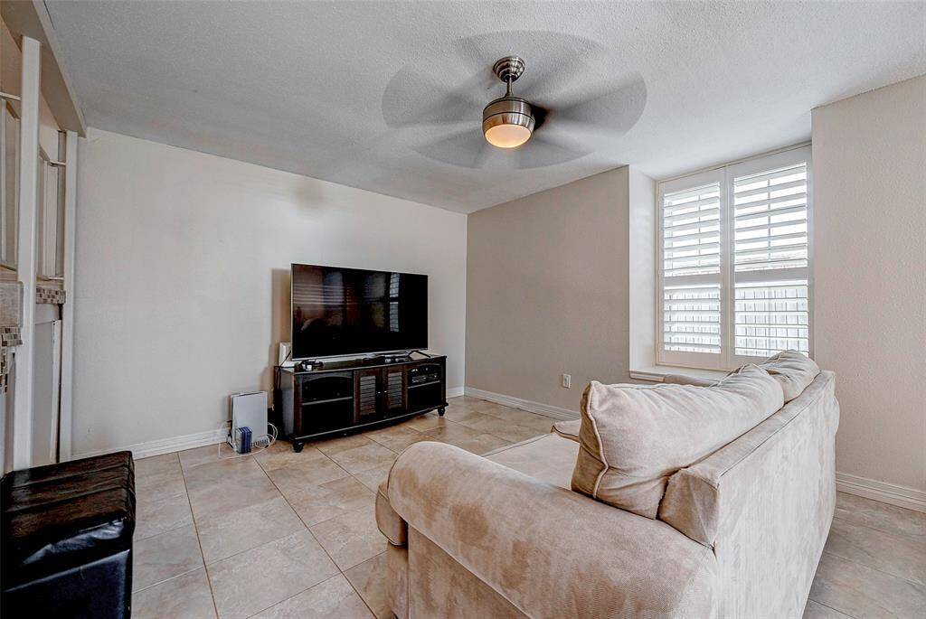 This cozy living room features a neutral color palette, tiled flooring, and a ceiling fan. It includes a large window with shutters for natural light and privacy.
