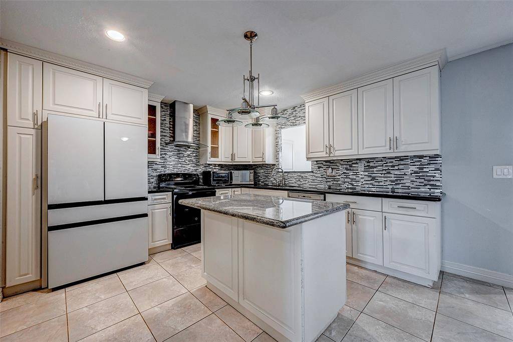 This kitchen features modern white cabinetry, a stylish tile backsplash, and a central island with a granite countertop. It includes stainless steel appliances, ample storage, and a sleek, contemporary light fixture, creating a bright and inviting cooking space.