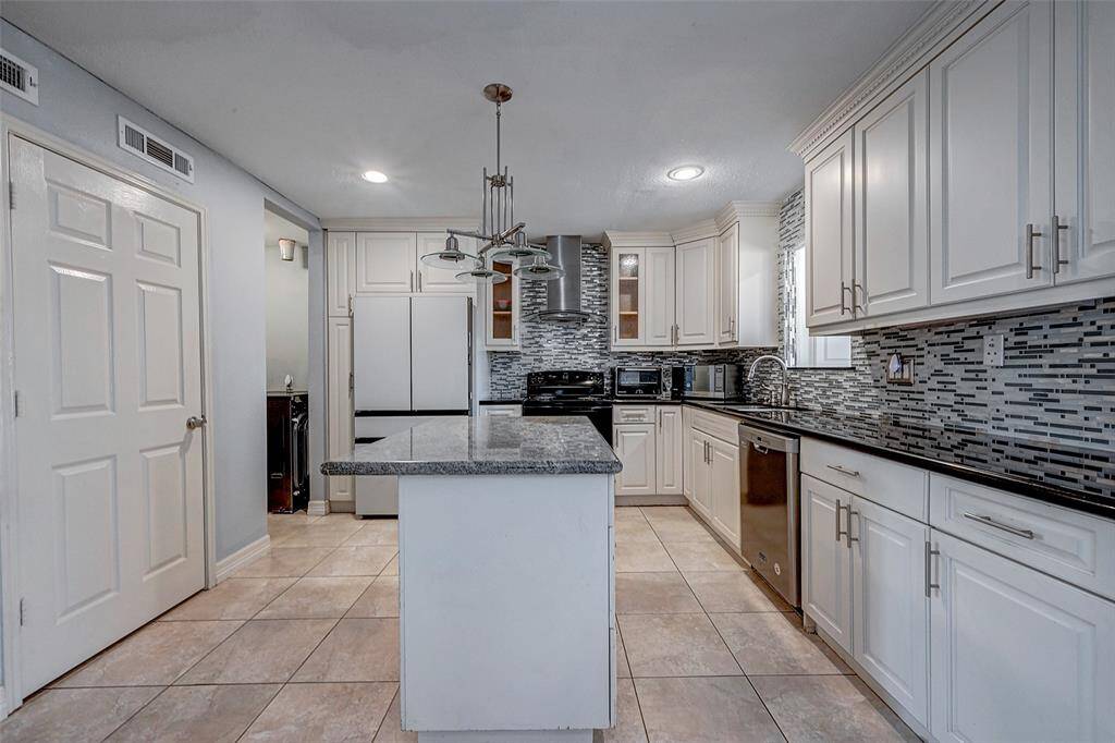This modern kitchen features sleek white cabinetry, a stylish tile backsplash, and a spacious island with granite countertops. Stainless steel appliances and ample lighting add functionality and elegance to the space.