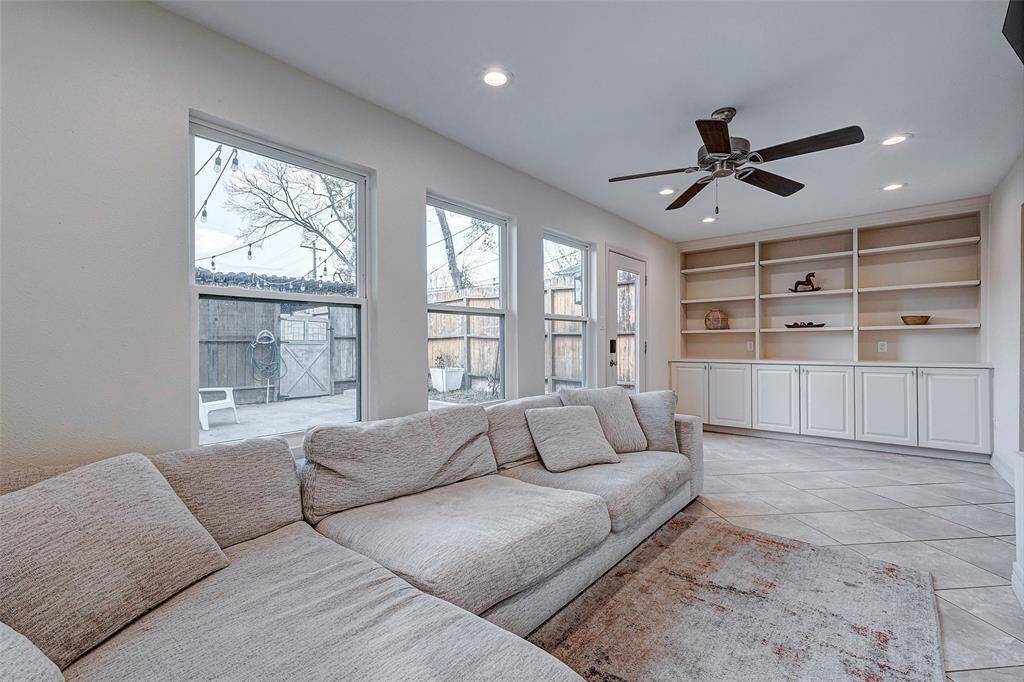 This cozy living room addition features, built-in shelving with cabinets, and a ceiling fan. Natural light streams in through multiple windows, offering a view of the backyard. The room is finished with neutral tiles and recessed lighting.