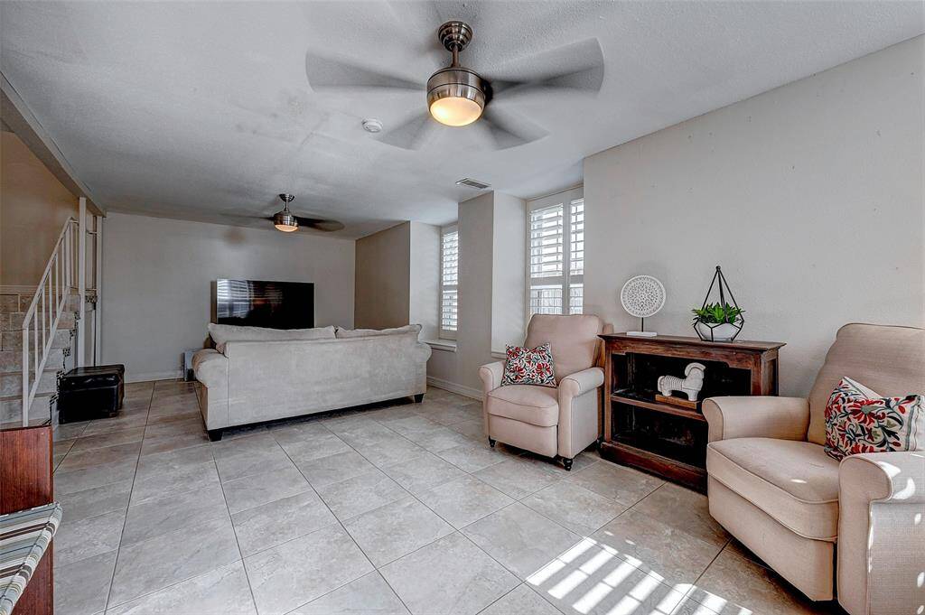 Spacious living room with ample natural light from large windows with shutters. Features modern ceiling fans and an open layout, perfect for relaxation and entertainment.