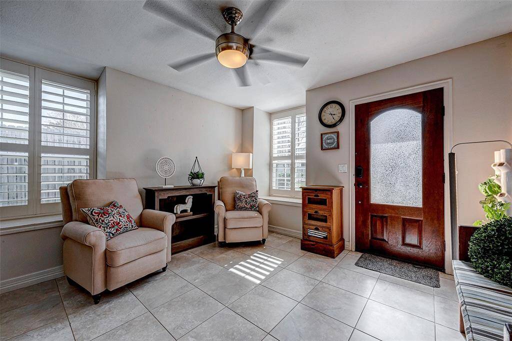 This cozy entryway features a warm wooden front door with frosted glass, tiled flooring, and ample natural light from large windows with shutters, creating an inviting atmosphere.