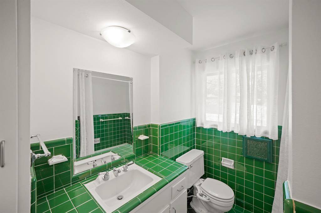 The Primary Bath offers a Retro Emerald Green Tile Topped Vanity, White Porcelain Undermount Sink, Polished Chrome Faucet and Handles, White Painted Cabinets and Drawers, Beveled Mirror, and Flush Mount Light Fixture above the Sink.