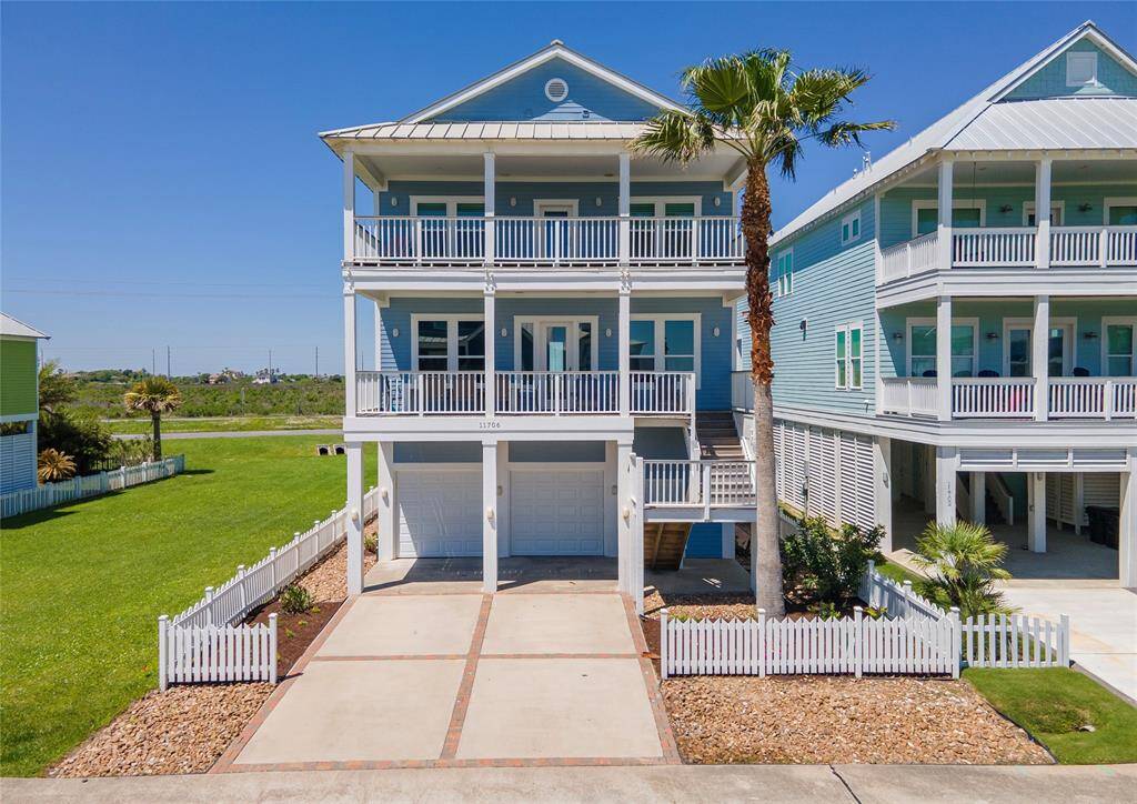 This photo showcases a modern, multi-story beach house with a light blue exterior and white trim. It features spacious balconies on each level, a large driveway leading to a double garage, and a neatly landscaped yard with palm trees and a white picket fence. Ideal for coastal living, it offers a bright and welcoming appearance.