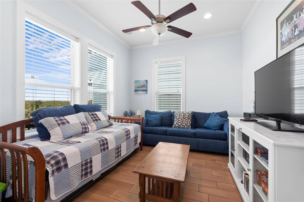 This cozy living room features a daybed with a quilt, a comfortable blue sofa, a wooden coffee table, and a TV on a white stand. Large windows and a ceiling fan enhance the bright, airy feel.