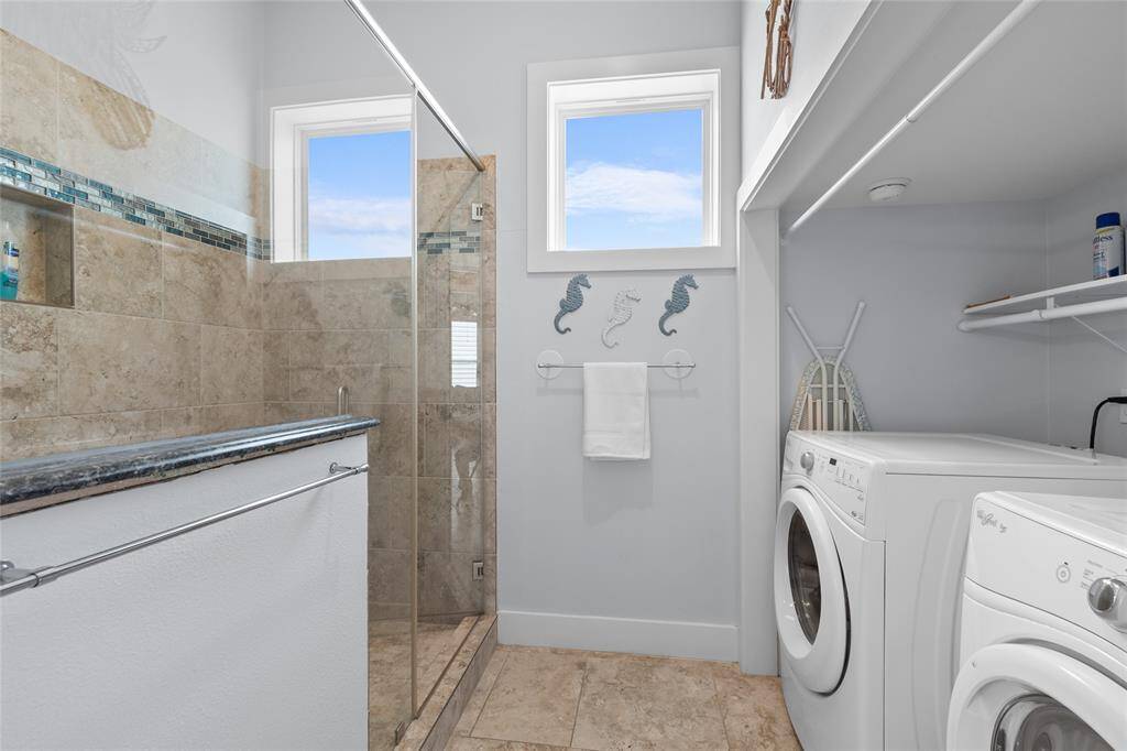 This photo showcases a combined bathroom and laundry space. It features a modern, glass-enclosed shower with tile accents, and a granite countertop. The laundry area includes a washer and dryer, with additional shelving for storage. Natural light enters through two small windows, enhancing the clean and functional design.